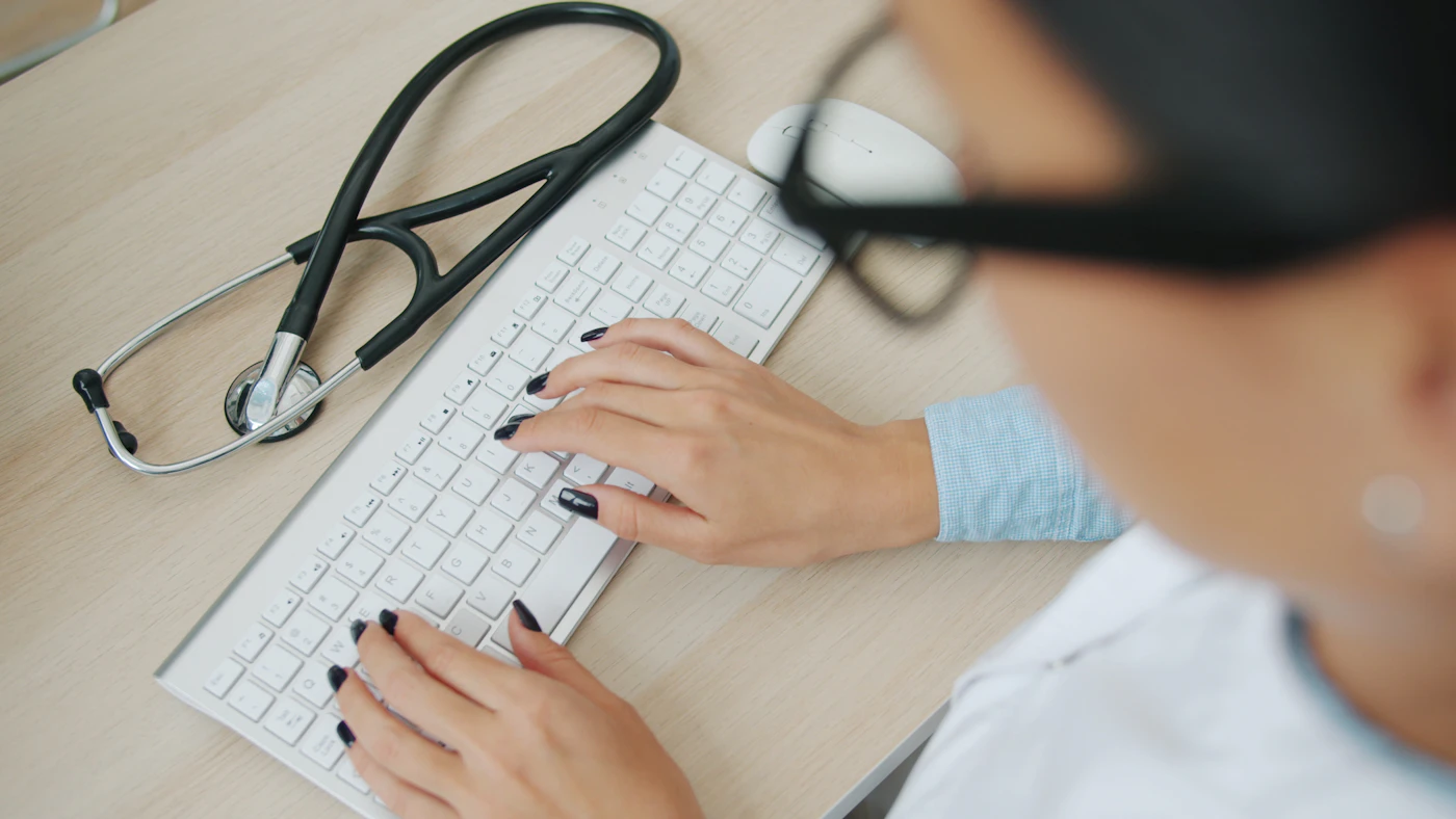 Physician typing visit notes on a keyboard with a stethoscope on the desk