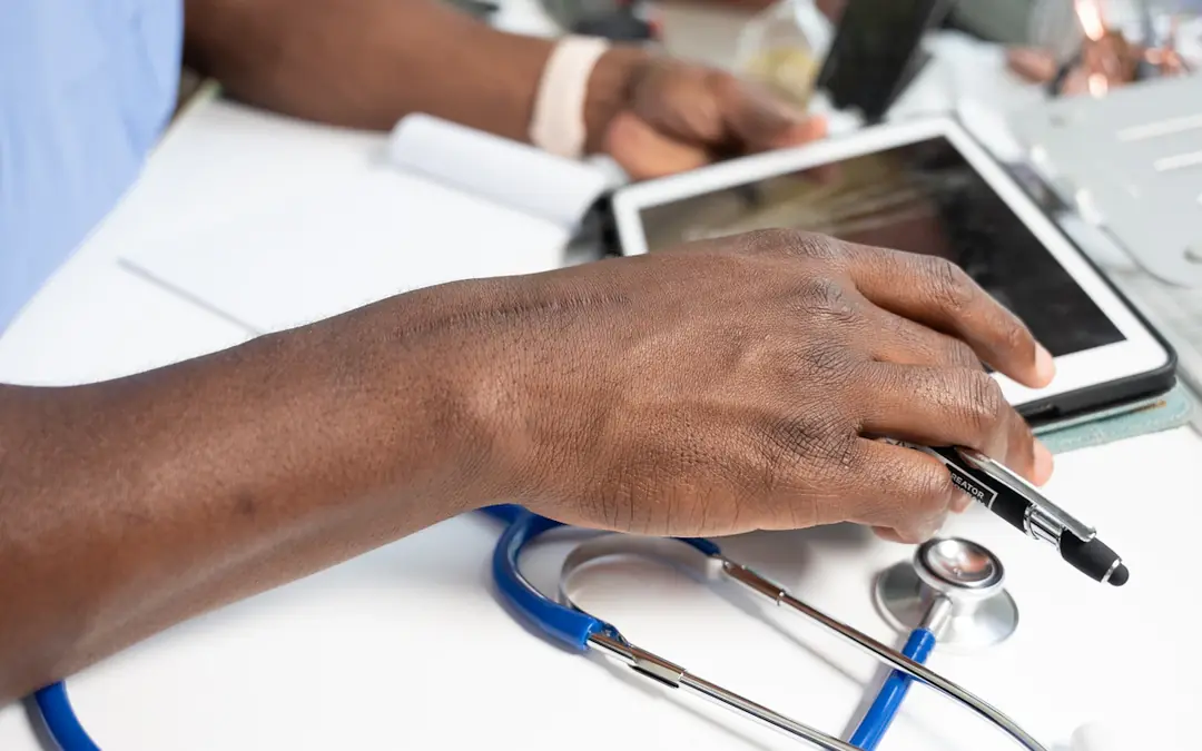 Hand holding a pen over a tablet and patient paperwork beside a stethoscope