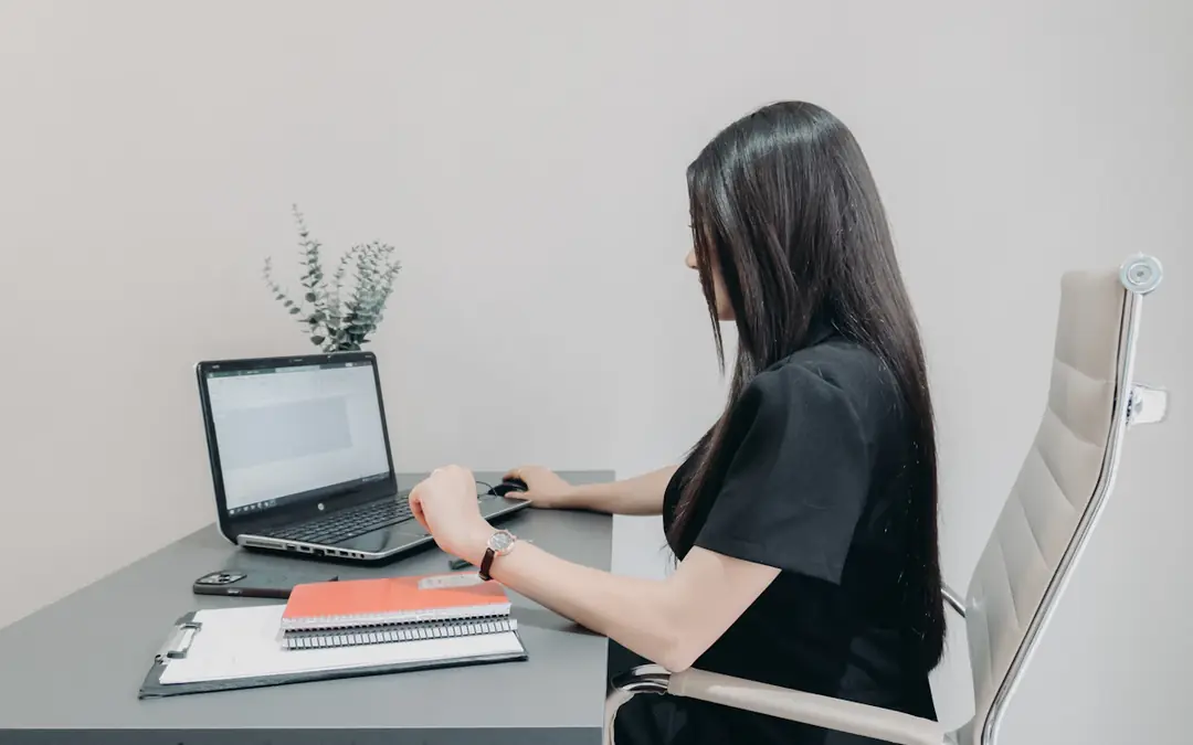 Woman working on a laptop at a desk with notebooks and a clipboard